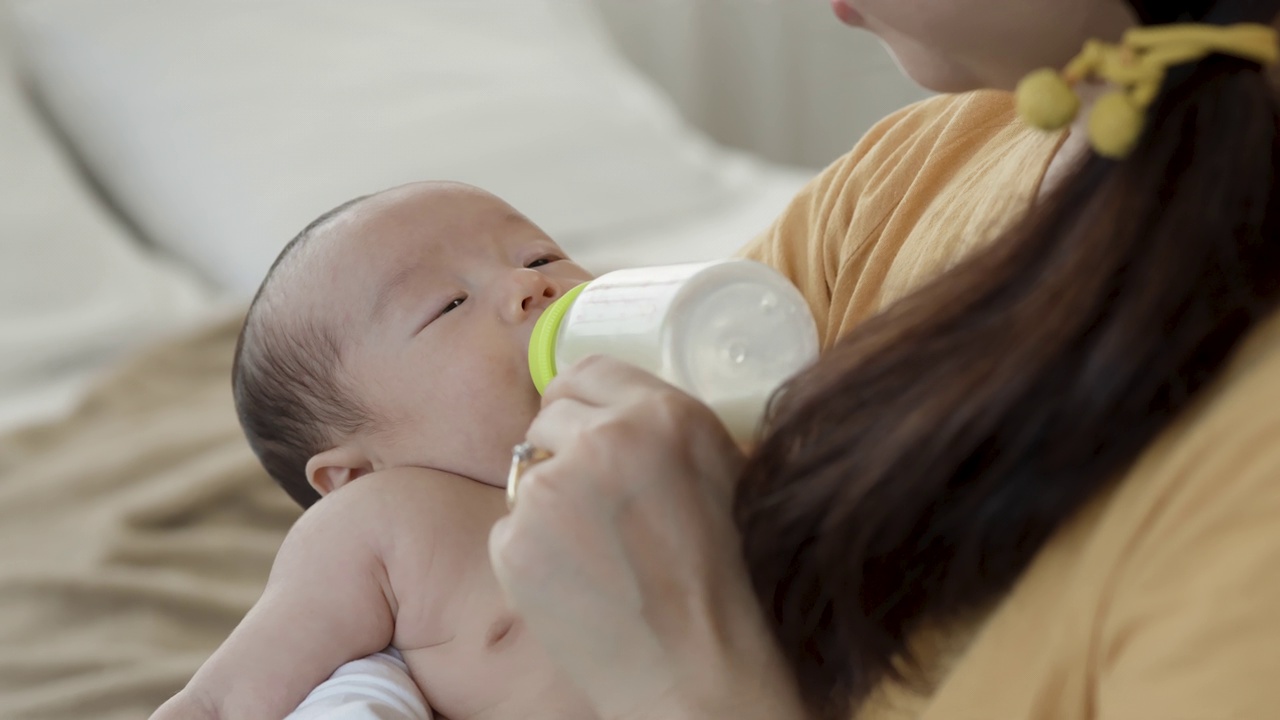 4k，一个两个月大的亚洲新生儿正在睡觉，吮吸着放在妈妈腿上的奶瓶，妈妈的手握着奶瓶。视频素材