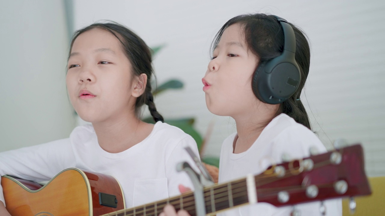 音乐教室里的快乐时光，7岁的亚洲小女孩弹吉他，5岁的亚洲小女孩戴着无线耳机，热情地唱歌。视频下载
