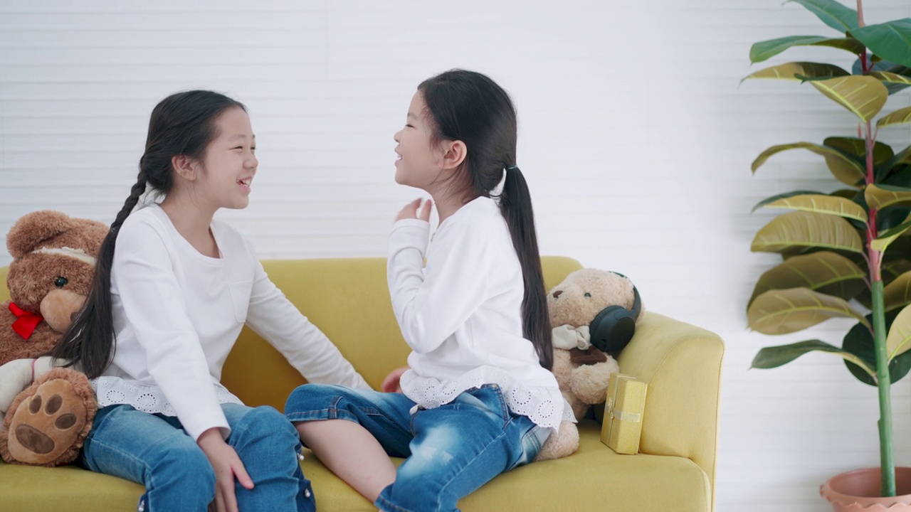 两个上小学的亚洲女孩坐在家里的沙发上一起唱歌。多年龄段的孩子或女性朋友们穿着白色t恤在上课前一起唱歌，兴奋地开始学习。快乐的时间。视频下载