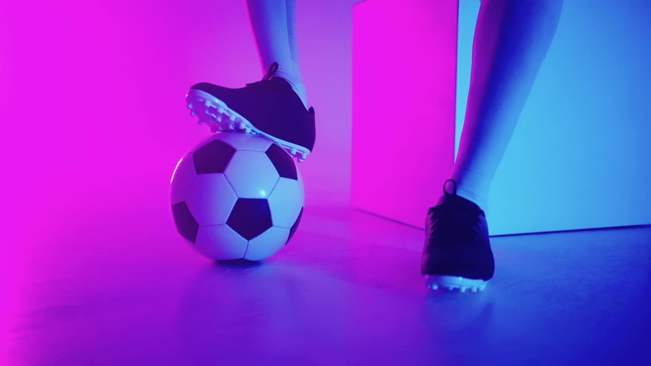在工作室的蓝红色霓虹灯下，一个职业黑人足球运动员的脚站在球上的慢镜头特写。巴西足球运动员脚踩球摆姿势视频下载