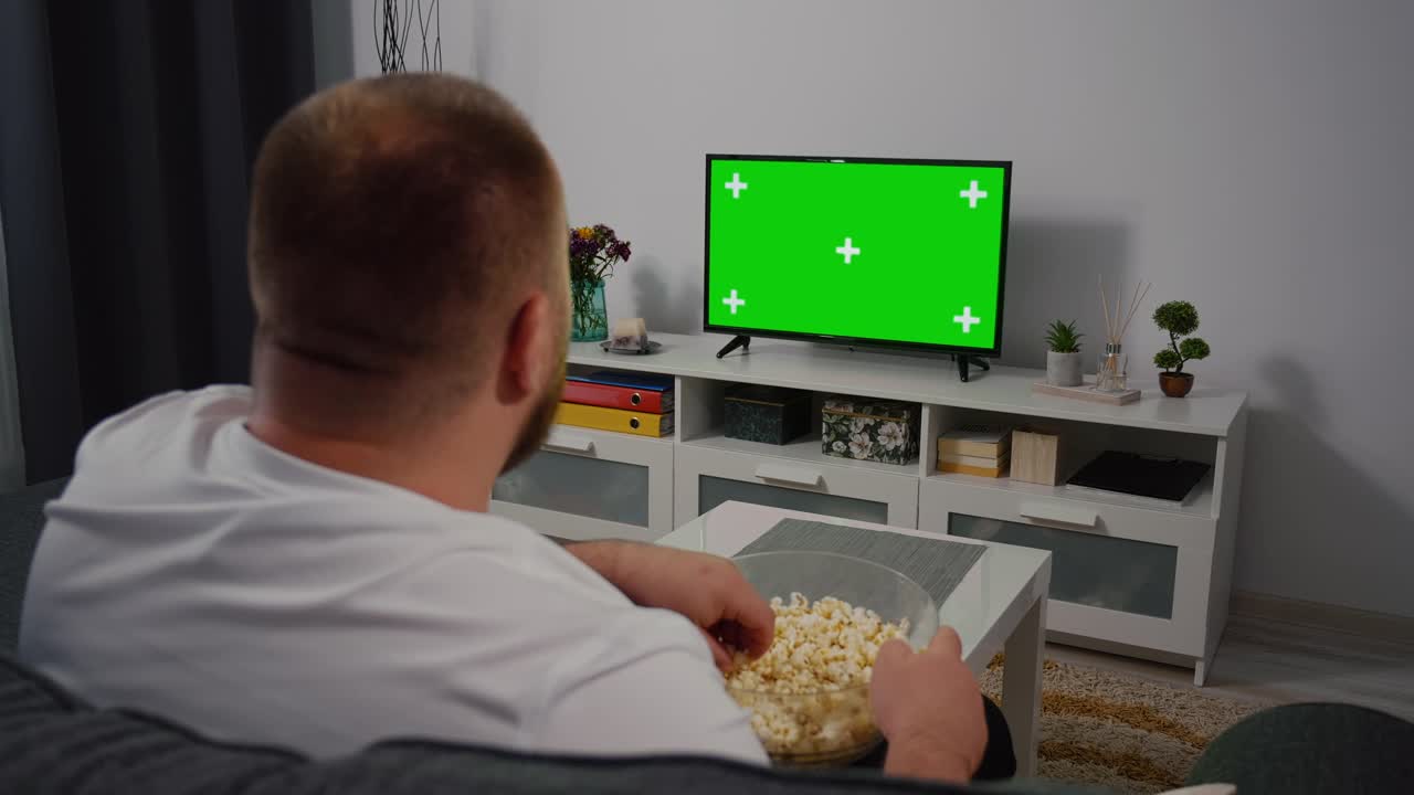 男人坐在家里的沙发上看绿色模拟屏幕电视。视频素材