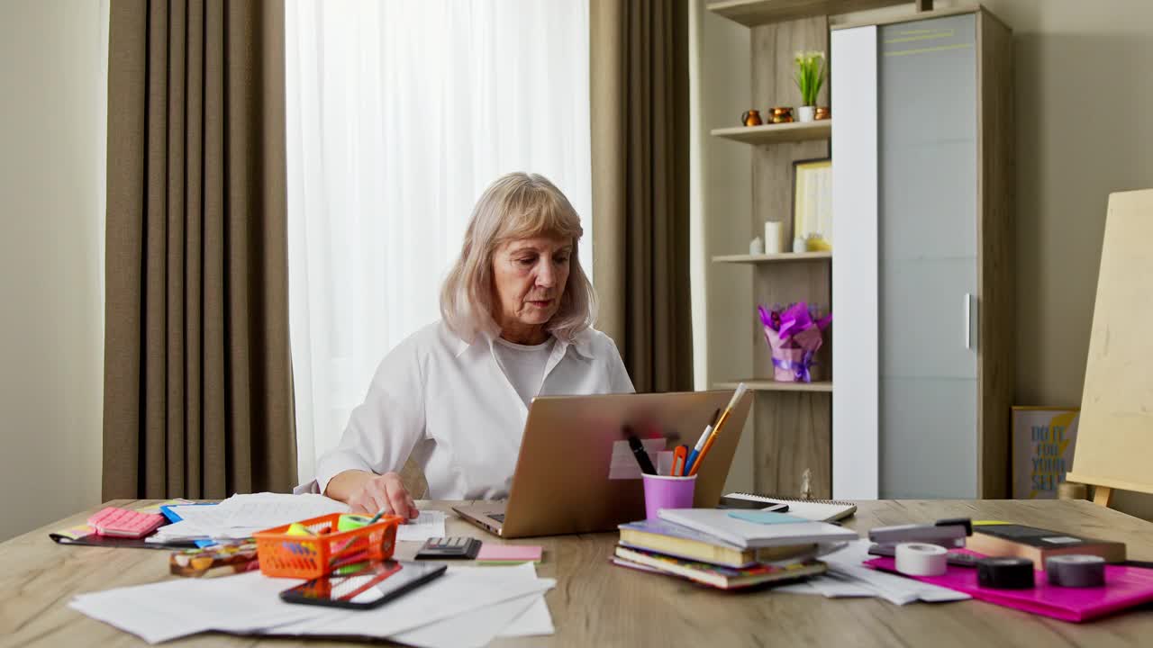 在Covid-19或冠状病毒大流行期间的封锁期间，年长的女商人在家在线工作，使用智能手机处理文书工作视频素材