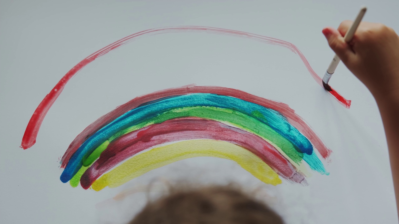 小女孩手里拿着画笔，用彩色颜料在白纸上画出一道彩虹。顶视图的片段视频素材