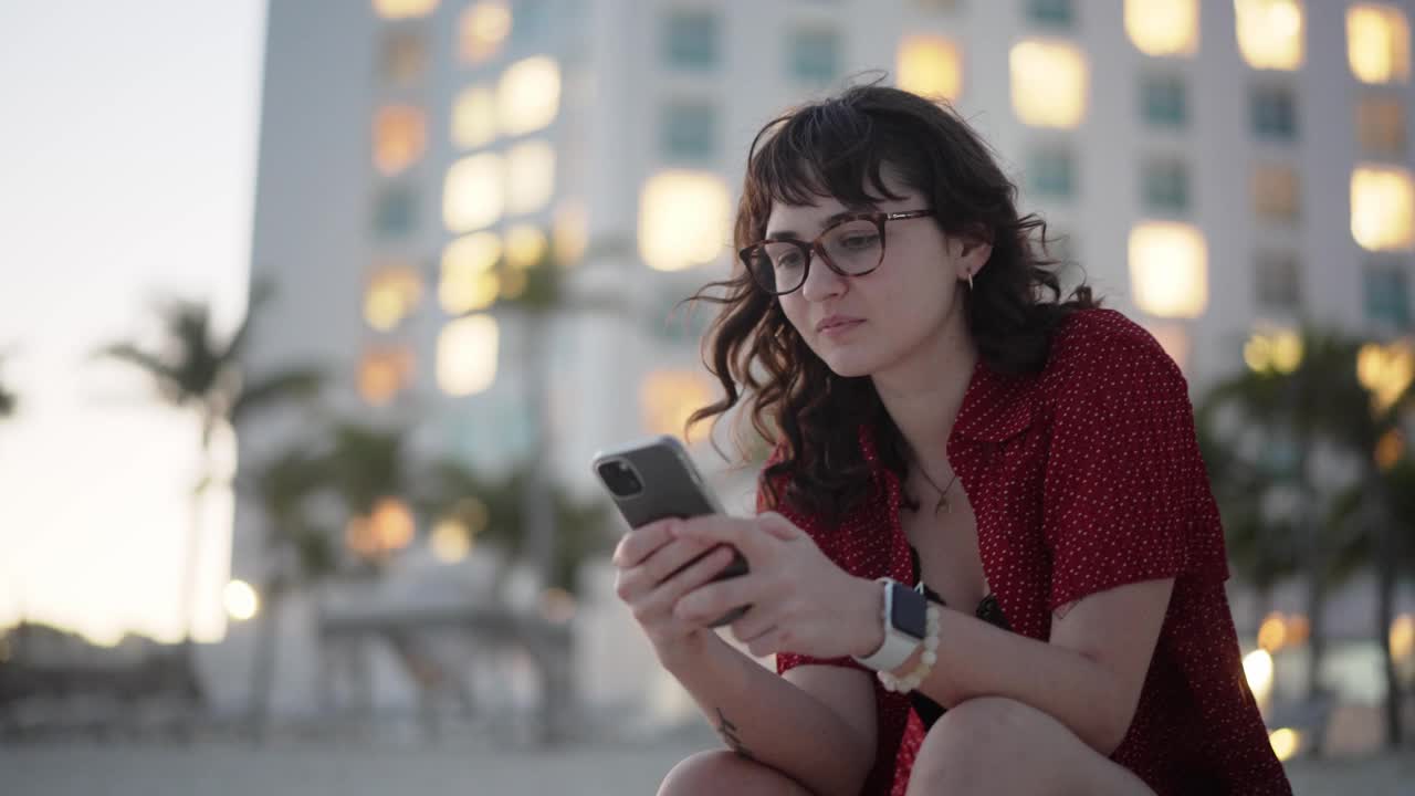 一名女子坐在酒店附近的海滩上用智能手机进行视频通话视频下载