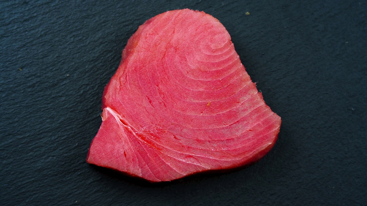 在黑石背景上旋转的金枪鱼片。生金枪鱼排是世界上最适合食用的鱼类。一块心形的鱼视频素材