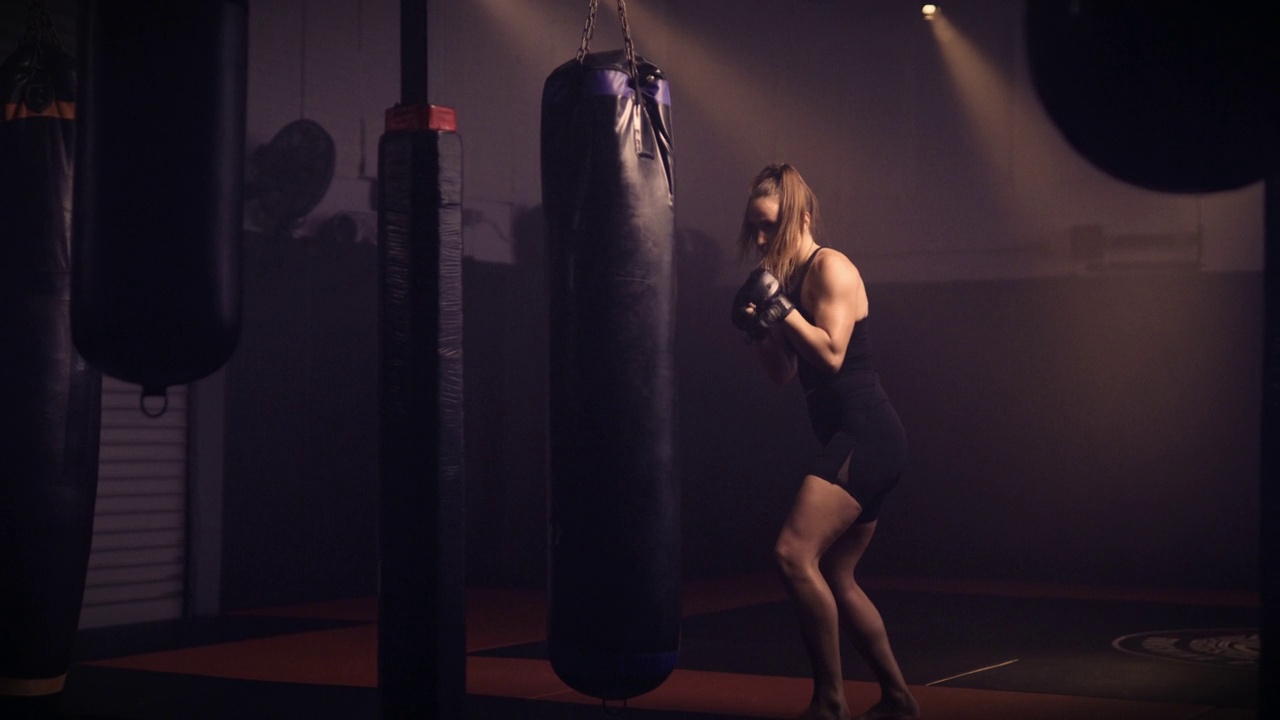 高端广角慢动作镜头的精英和强大的综合武术女战士，跆拳道和拳击沉重的拳击袋。背景设置在一个忧郁的综合格斗/拳击馆。低调的照明视频下载