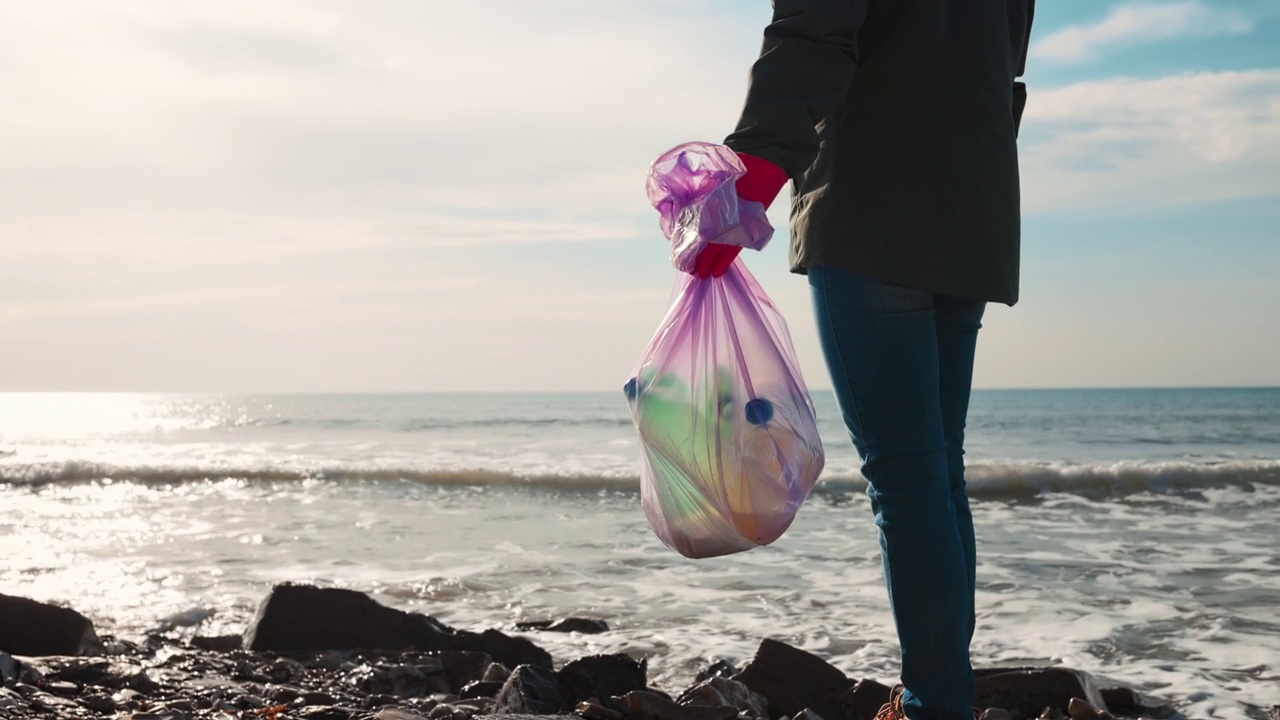 一位女性志愿者站在海边，手里拿着一个装着塑料瓶的垃圾袋。缩小。世界环境日与生态的概念视频素材
