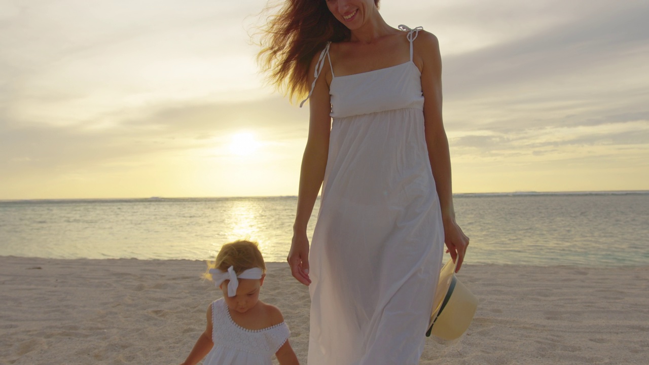 在慢镜头中，小女孩和妈妈在热带海滩散步。夕阳在背景中视频下载