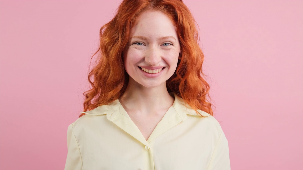 一个积极的红发年轻女子站在粉色背景的摄影棚里，孤立地站在镜头前大笑。视频下载
