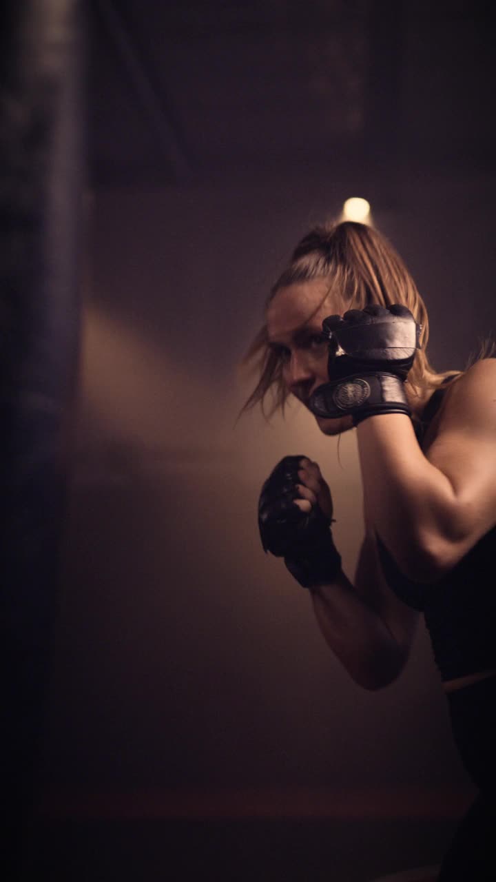 高端慢动作镜头的精英和强大的综合武术女战士，跆拳道和拳击沉重的拳击袋。在垂直/移动视频长宽比和设置在一个穆迪的综合格斗/拳击健身房。低调的照明视频下载