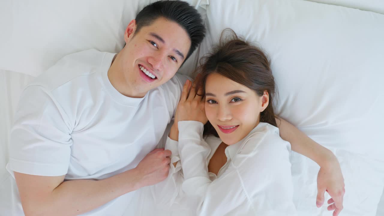 亚洲新婚夫妇躺在床上看着相机的肖像。漂亮漂亮的年轻男女穿着睡衣在家里的卧室里享受清晨的活动。家庭关系的概念视频素材