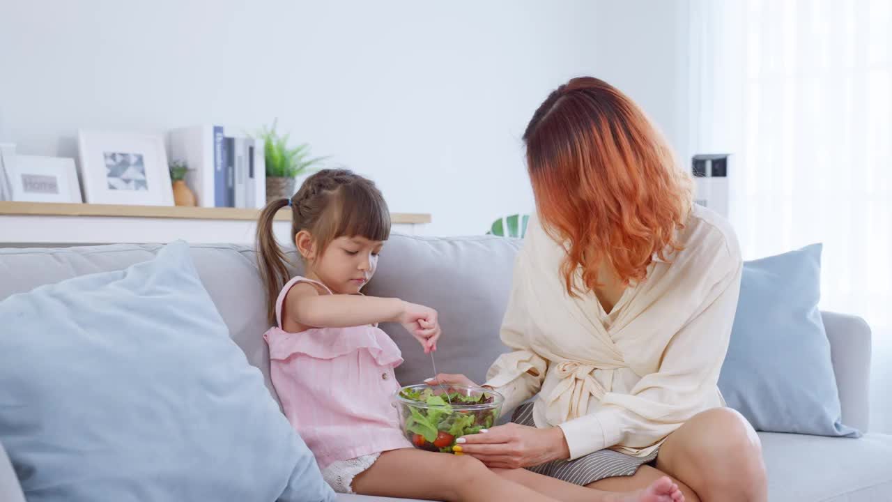 亚洲幸福家庭，父母和孩子一起在家度过空闲时间。快乐的小女孩和妈妈坐在客厅的沙发上，吃着蔬菜沙拉。内部活动关系。视频素材