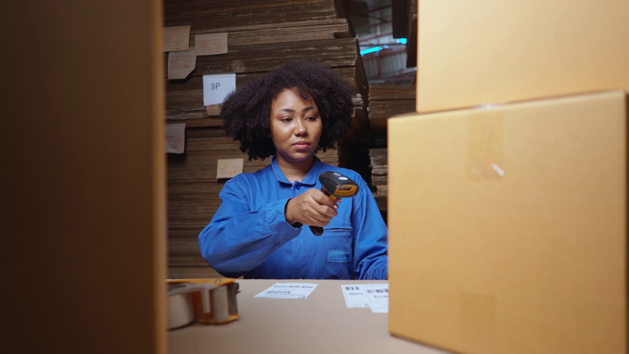 身着蓝色连体衣的非洲年轻女性在物流公司仓库检查标签，准备将产品送到客户手中，检查清单，使用条形码扫描仪，用胶带包装箱子视频素材