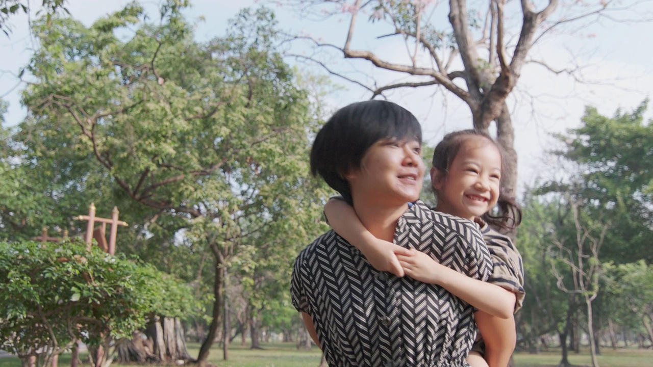 亚洲母亲正在玩，背着她可爱的女儿在美丽的公园。它表达了爱和关心的关系与幸福和快乐，这对孩子是有好处的。视频购买
