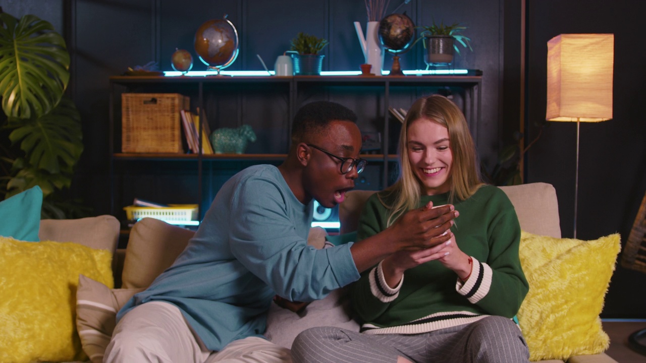 快乐的年轻白人金发女子使用手机时感到惊讶和惊讶，与黑人男性朋友分享。视频下载
