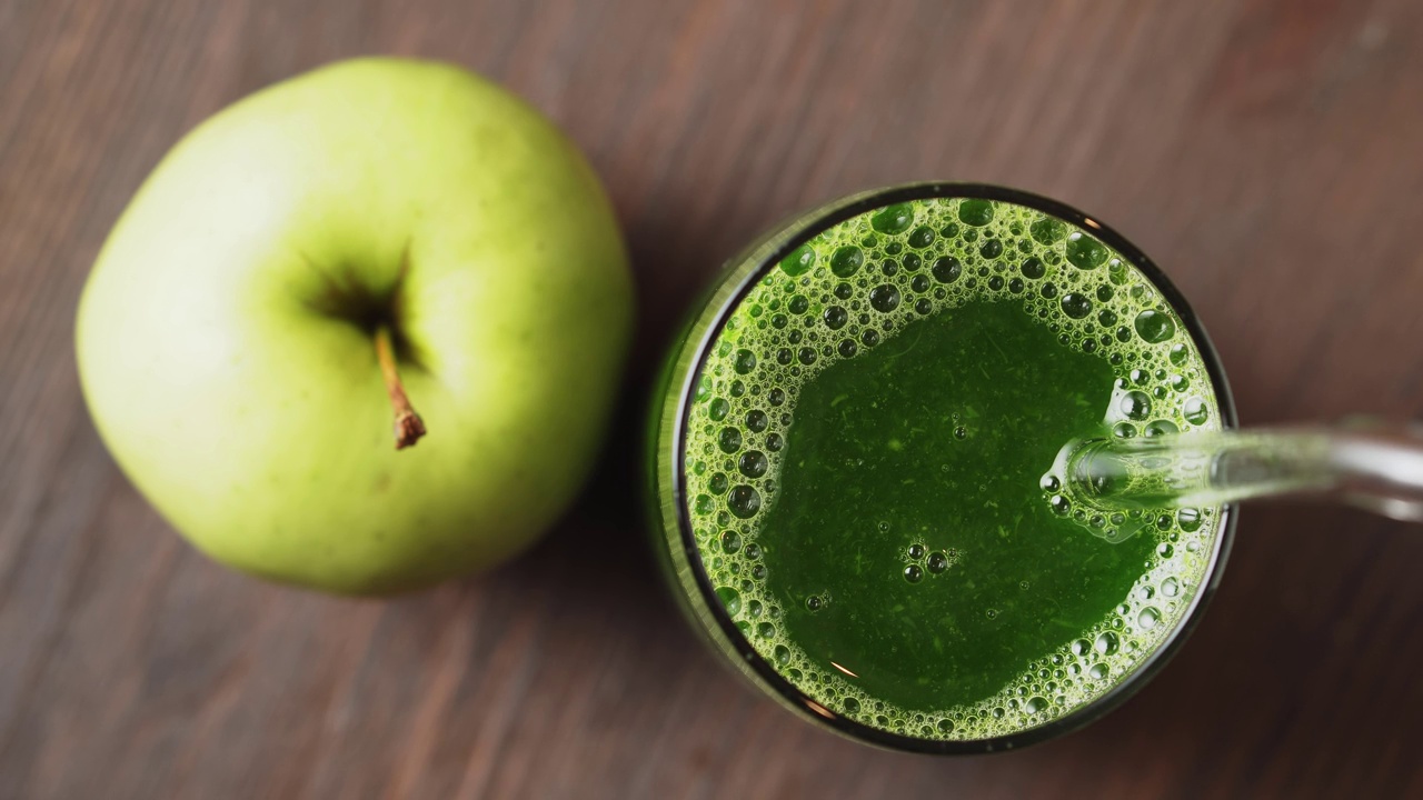 美味的绿色冰沙在玻璃杯中，与饮料吸管混合，木制背景，俯视图。健康饮食的概念。视频下载