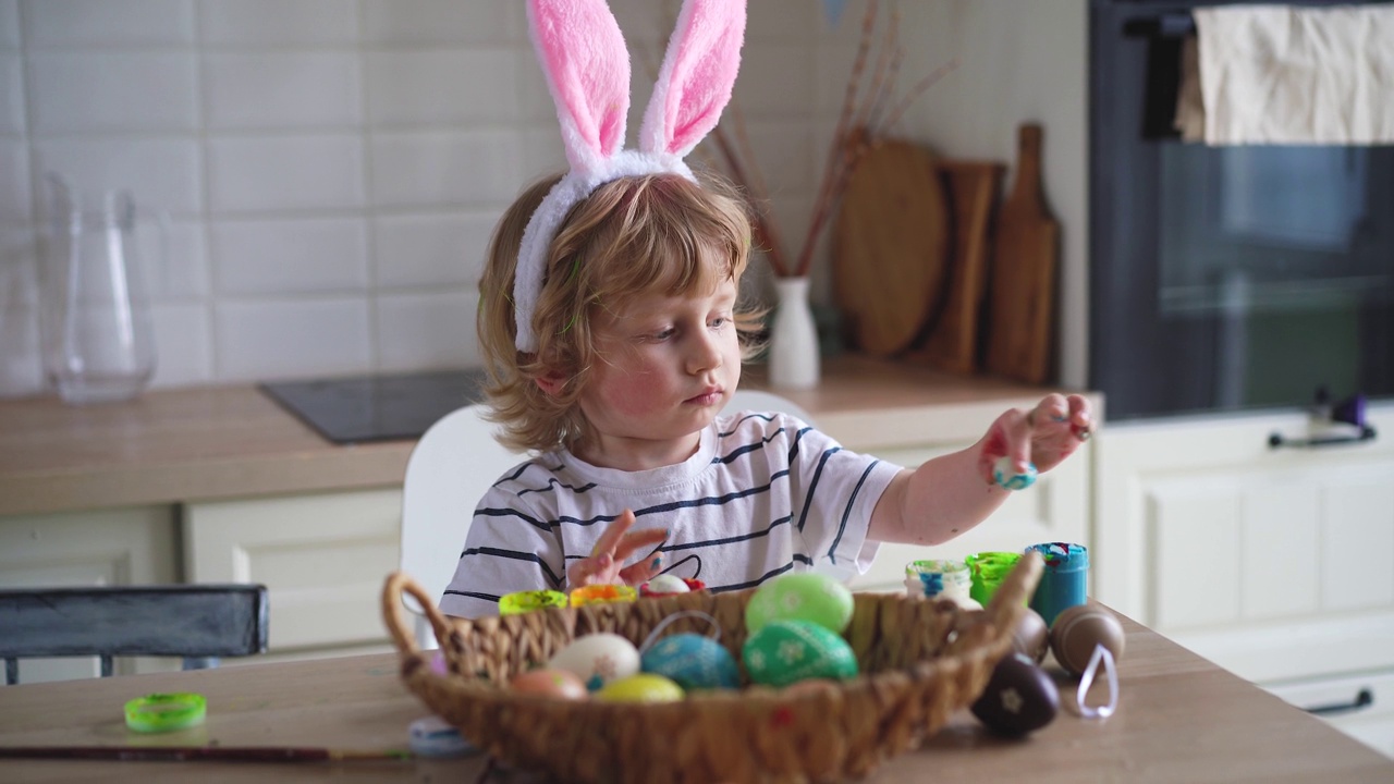 可爱的两岁小男孩戴着兔子耳朵，坐在厨房的桌子旁，用五颜六色的颜料画复活节彩蛋视频下载