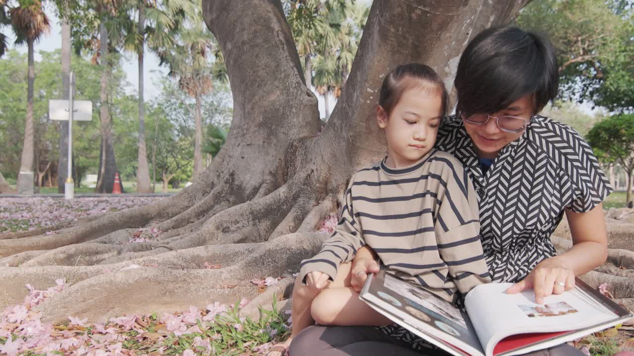一位亚洲妈妈和她的女儿在美丽的公园里的一棵树下看书。它体现了父母关爱和家庭学习的理念，这是儿童户外生活方式的休闲活动。视频下载