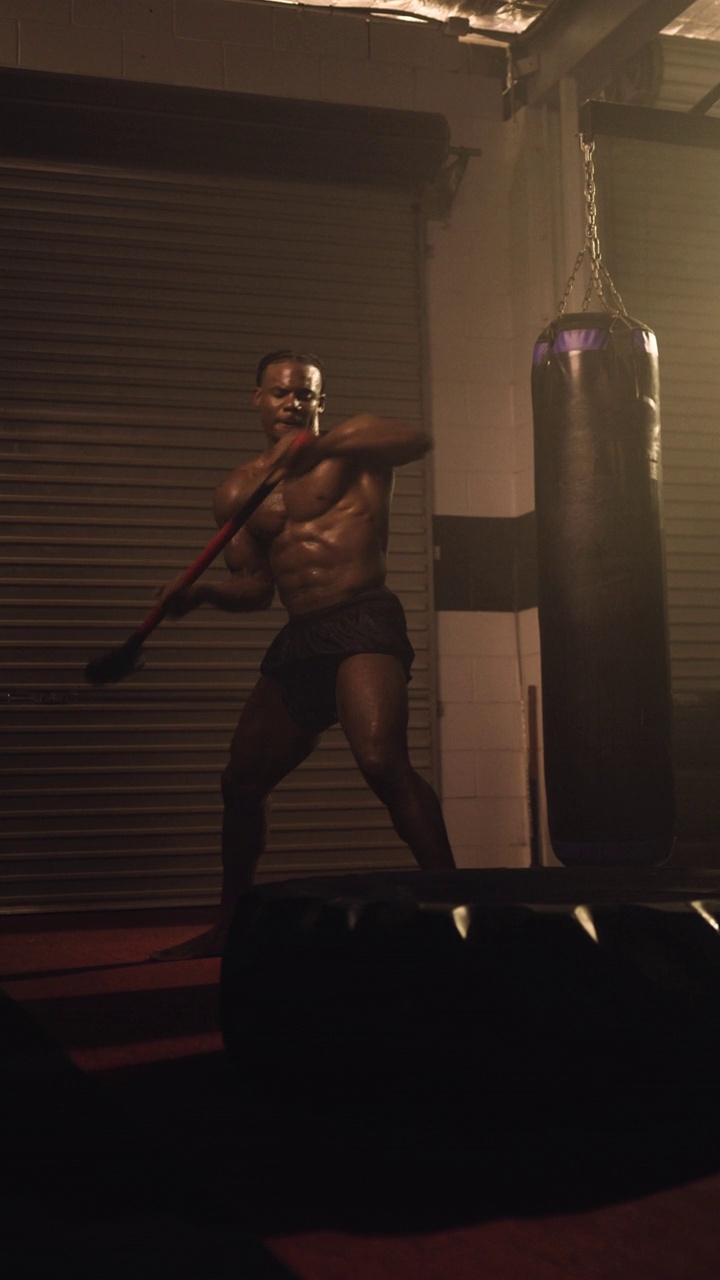 一个高端垂直移动视频拍摄的精英非裔美国健美运动员，表演一个大锤打击拖拉机轮胎/轮胎。设置在一个穆迪，低关键照明混合武术体育馆与烟雾的散射视频素材