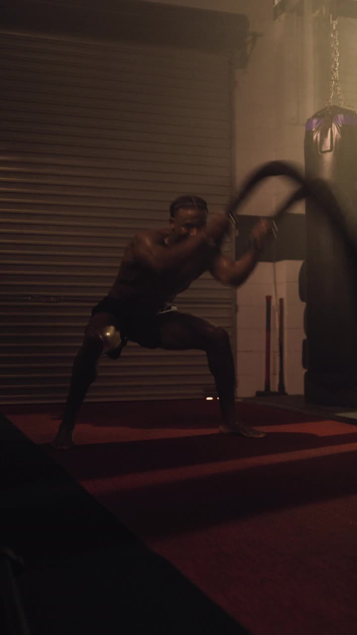 一个高端垂直移动视频拍摄的精英非裔美国健美运动员，执行一个重量级交替波浪战斗绳运动。设置在一个穆迪，低关键照明混合武术体育馆与烟雾的散射视频素材