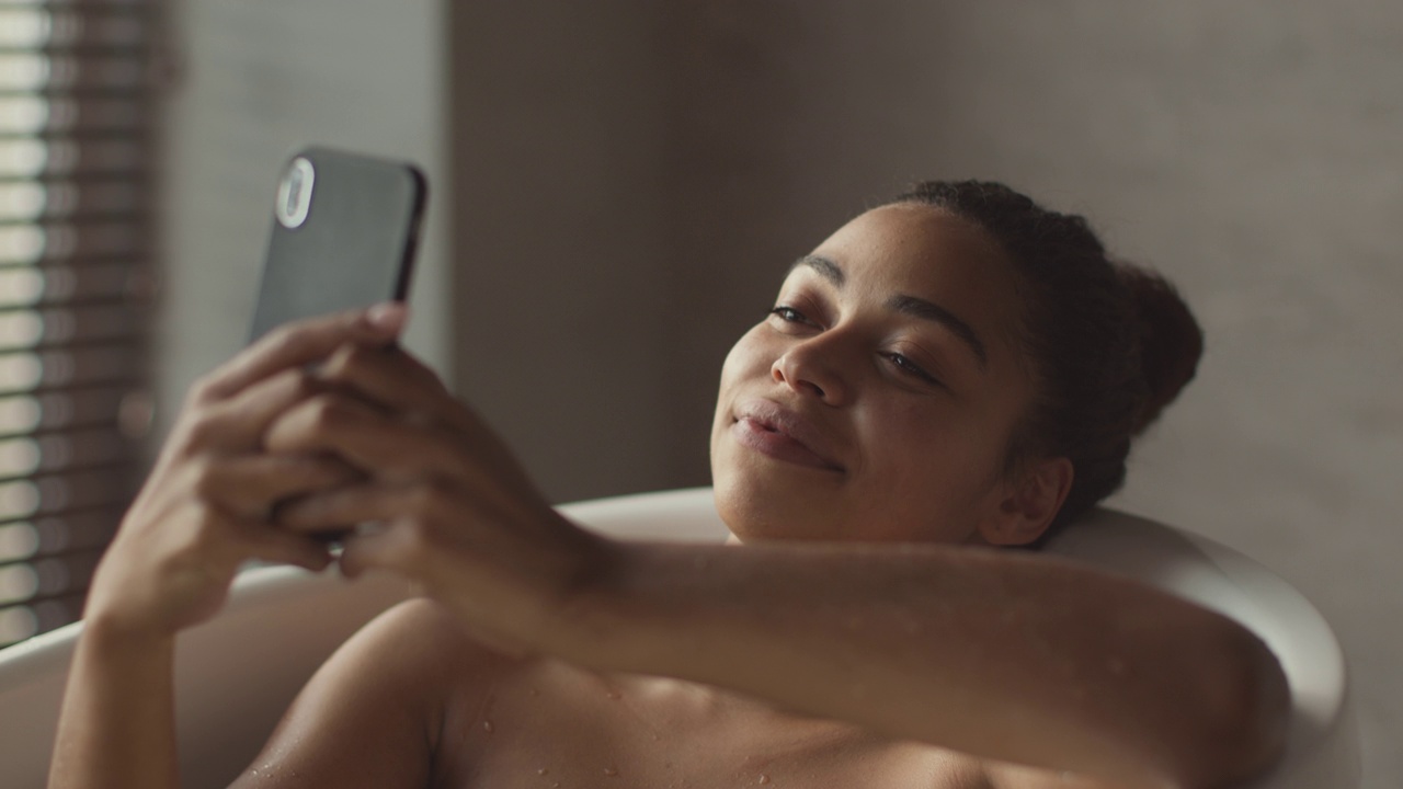 年轻可爱的非洲裔美国女士放松在浴室在家里，在智能手机上上网社交媒体，跟踪拍摄视频下载