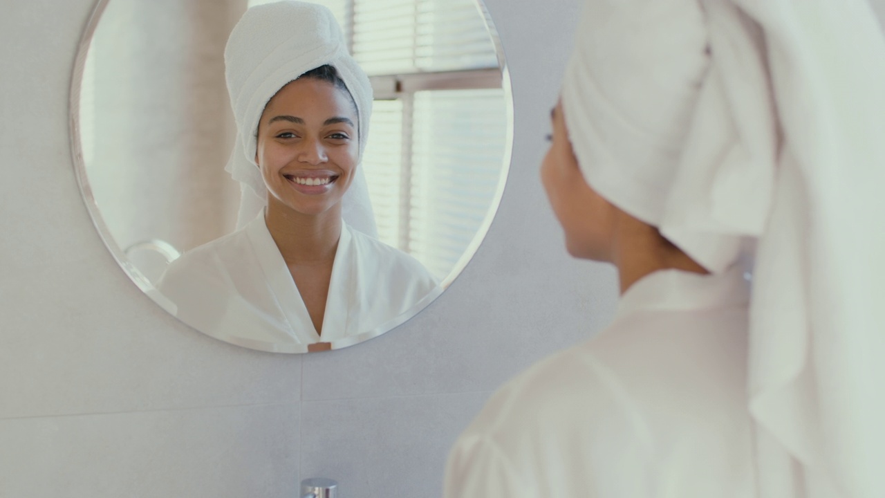 年轻的非洲裔美国妇女穿着浴袍，头戴毛巾，淋浴后对着镜子微笑视频素材
