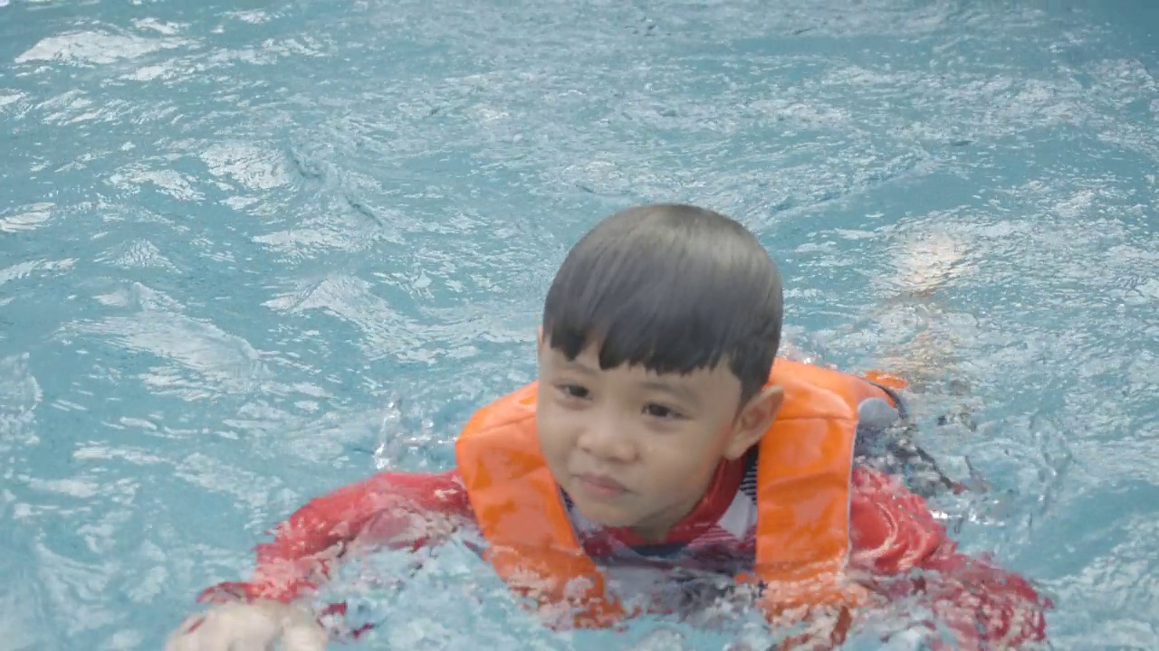 快乐的亚洲小男孩穿着泳装和救生衣，在游泳池里学习游泳和玩水，微笑着快乐和乐趣。暑假休闲活动或儿童假期旅游。视频素材