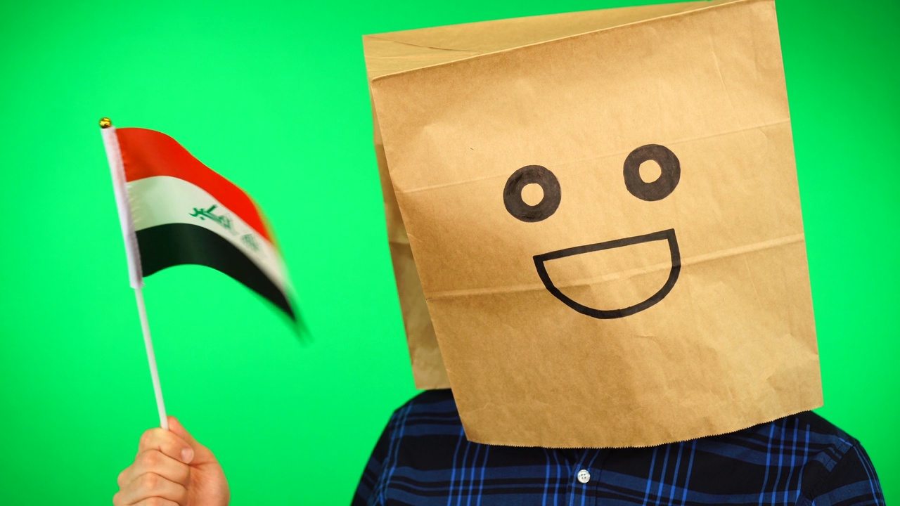 头上戴着纸袋的男子的肖像，在绿色的背景下挥舞着伊拉克国旗，面带微笑。视频素材