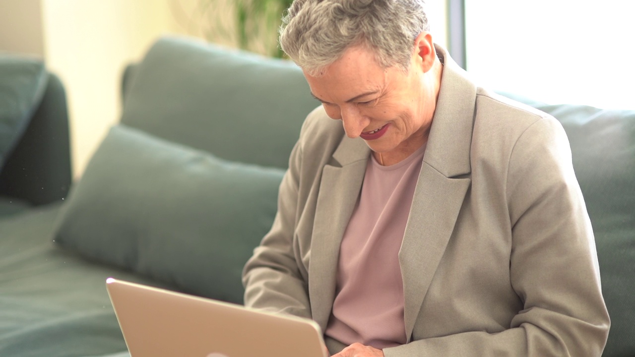 穿着西装、面带微笑的白发女士坐在沙发上独自用笔记本电脑工作。远程就业理念，女性自由职业者，幸福退休视频下载