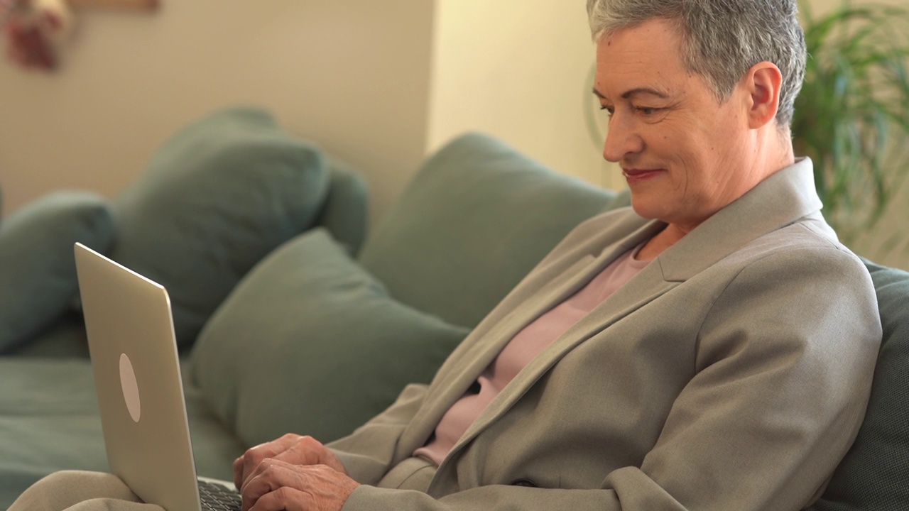 一头灰白短发的白人女性独自坐在沙发上，用笔记本电脑上网或购物视频下载