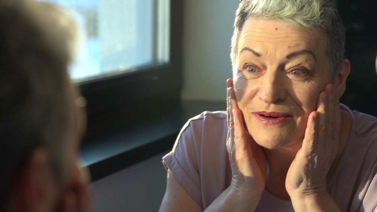 一个灰白头发的老年妇女的特写肖像，皮肤护理和化妆。有吸引力的资深女性看着自己在镜子里视频下载