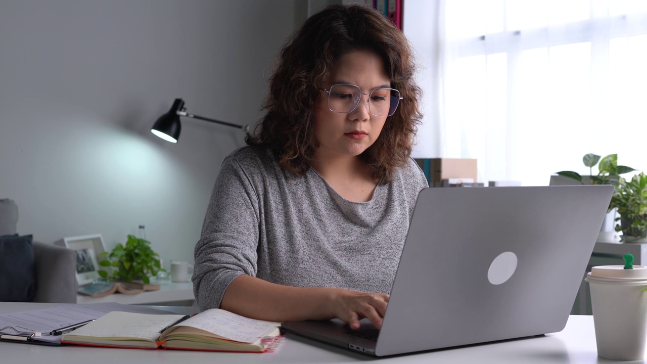 年轻的亚洲妇女坐在看起来筋疲力尽，在使用笔记本电脑在家庭办公室工作。忧心忡忡的亚洲妇女看着笔记本电脑，对电脑问题感到沮丧视频素材