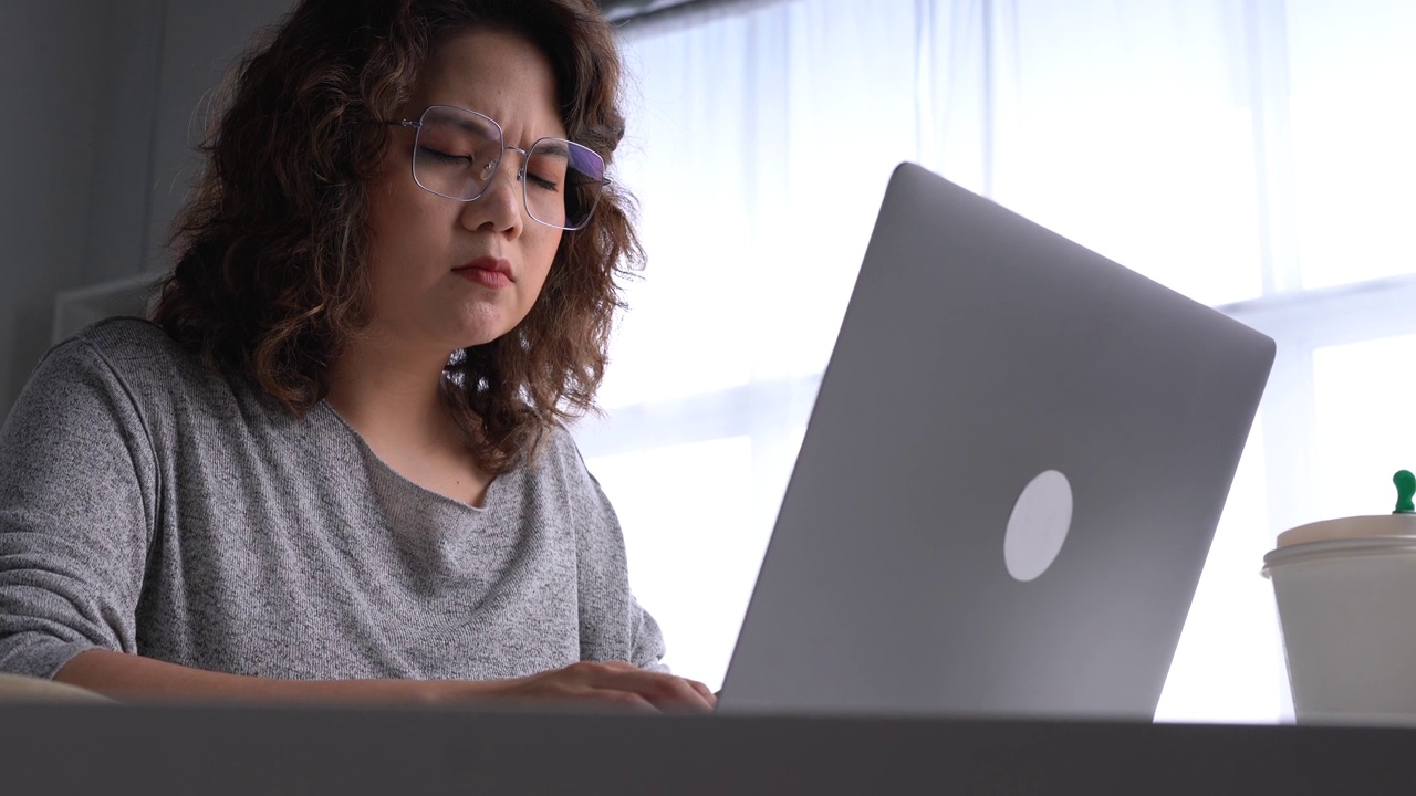 年轻的亚洲妇女坐在看起来筋疲力尽，在使用笔记本电脑在家庭办公室工作。忧心忡忡的亚洲妇女看着笔记本电脑，对电脑问题感到沮丧视频素材