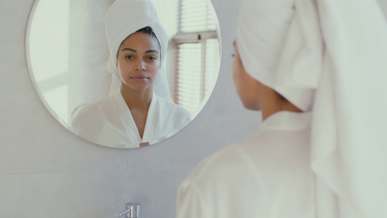 年轻和平的非裔美国妇女的肖像穿着浴衣和毛巾在头上看着浴室的镜子视频素材