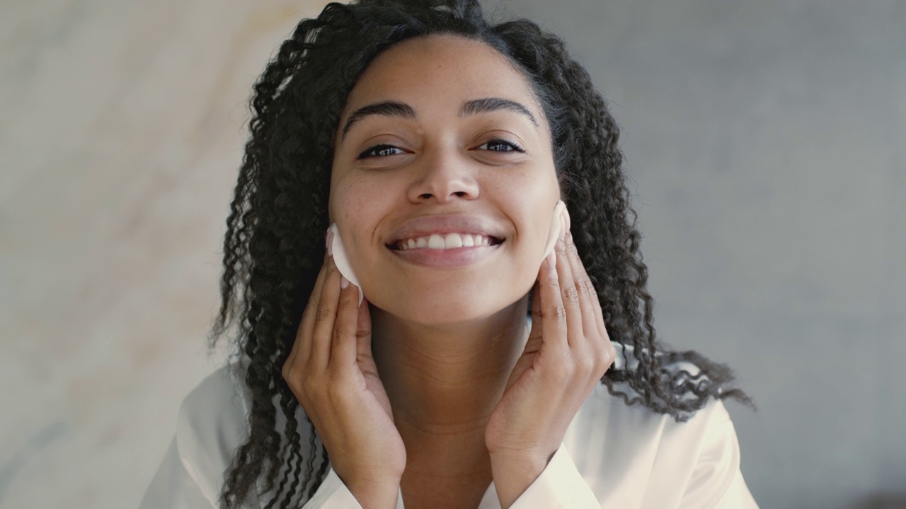 去除。快乐的非洲裔美国妇女清洁她的脸胶束水和两个棉垫，镜子pov视频素材