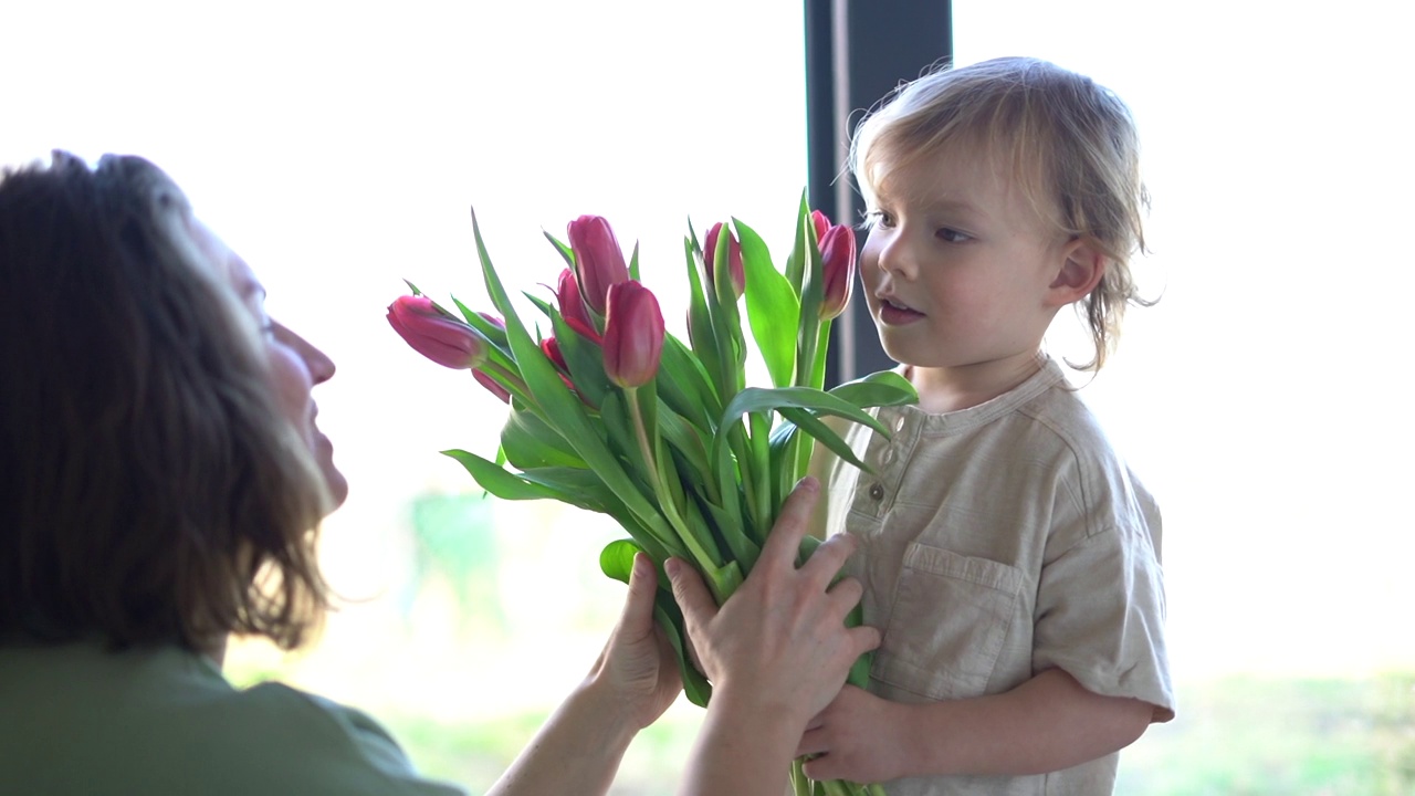 可爱的金发小男孩祝他年轻的妈妈生日快乐，并送给她一束红色的郁金香。春节和母亲节的概念。母亲和儿子视频下载