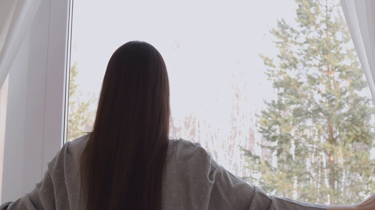清晨，一个深色头发的女人拉开窗帘，望向窗外一片雪中的针叶林。在乡间别墅休息。美好的早晨从第一缕阳光开始视频素材