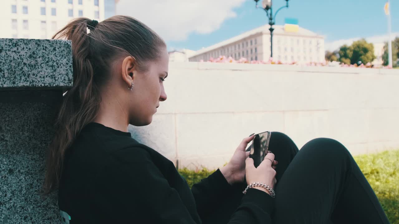 年轻女子坐在街道公园的栏杆上使用智能手机视频下载