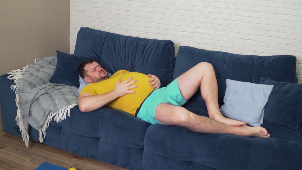 一个留着胡子、穿着黄色t恤的胖男人正躺在家里的床上，抚摸着他生病的大肚子。腹胀，暴饮暴食，痉挛，疼痛，腹泻。适当的营养视频素材