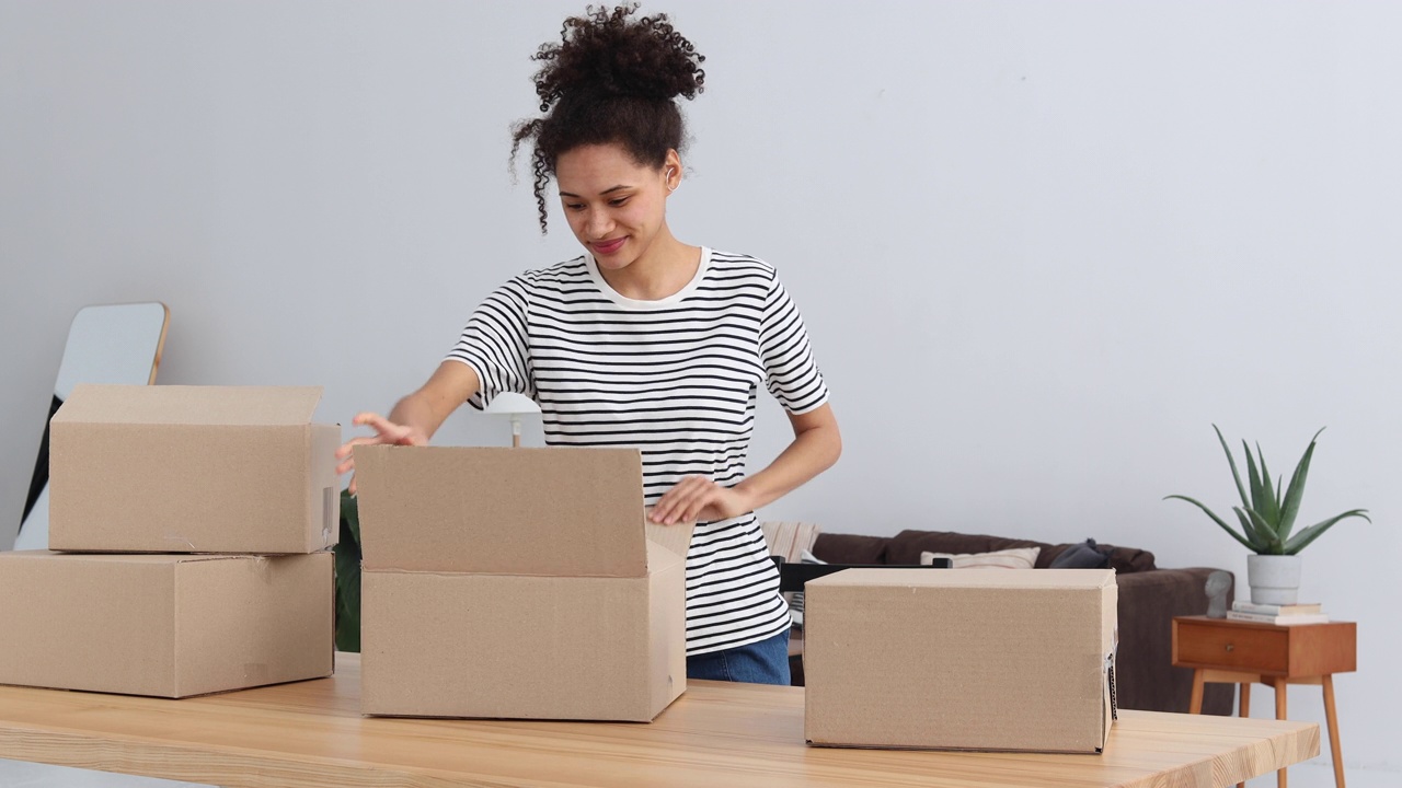 一名年轻的非洲裔美国妇女站在家里，打开一个装有从网上购买的衣服的箱子。包裹,交付的概念视频下载