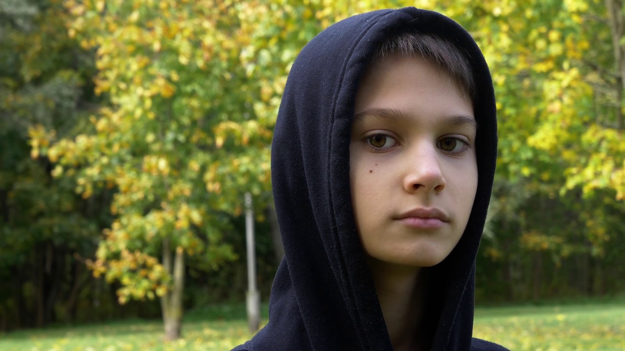 十几岁的男孩在户外穿着黑色连帽夹克的特写照片，背景是城市公园的树木。4 k录像视频视频下载