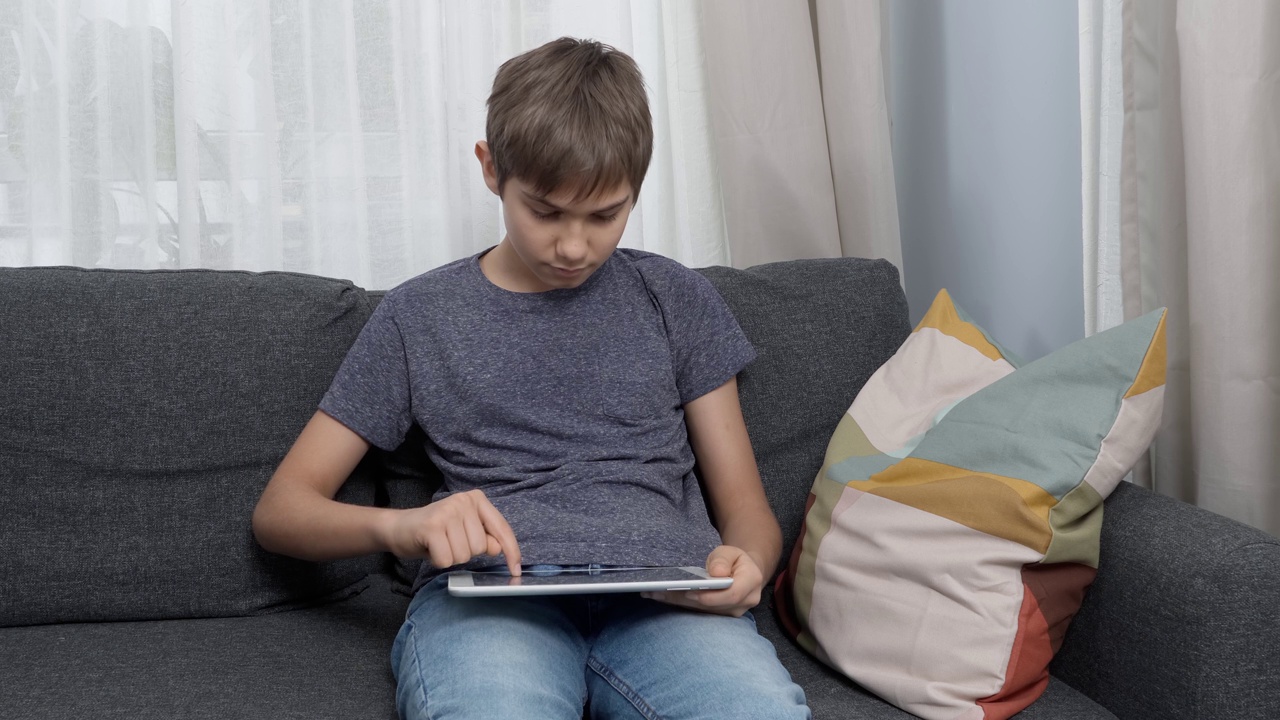 十几岁的男孩坐在家里的沙发上使用数码平板电脑。孩子使用应用程序学习，玩游戏，上网，看视频，在线学习视频素材