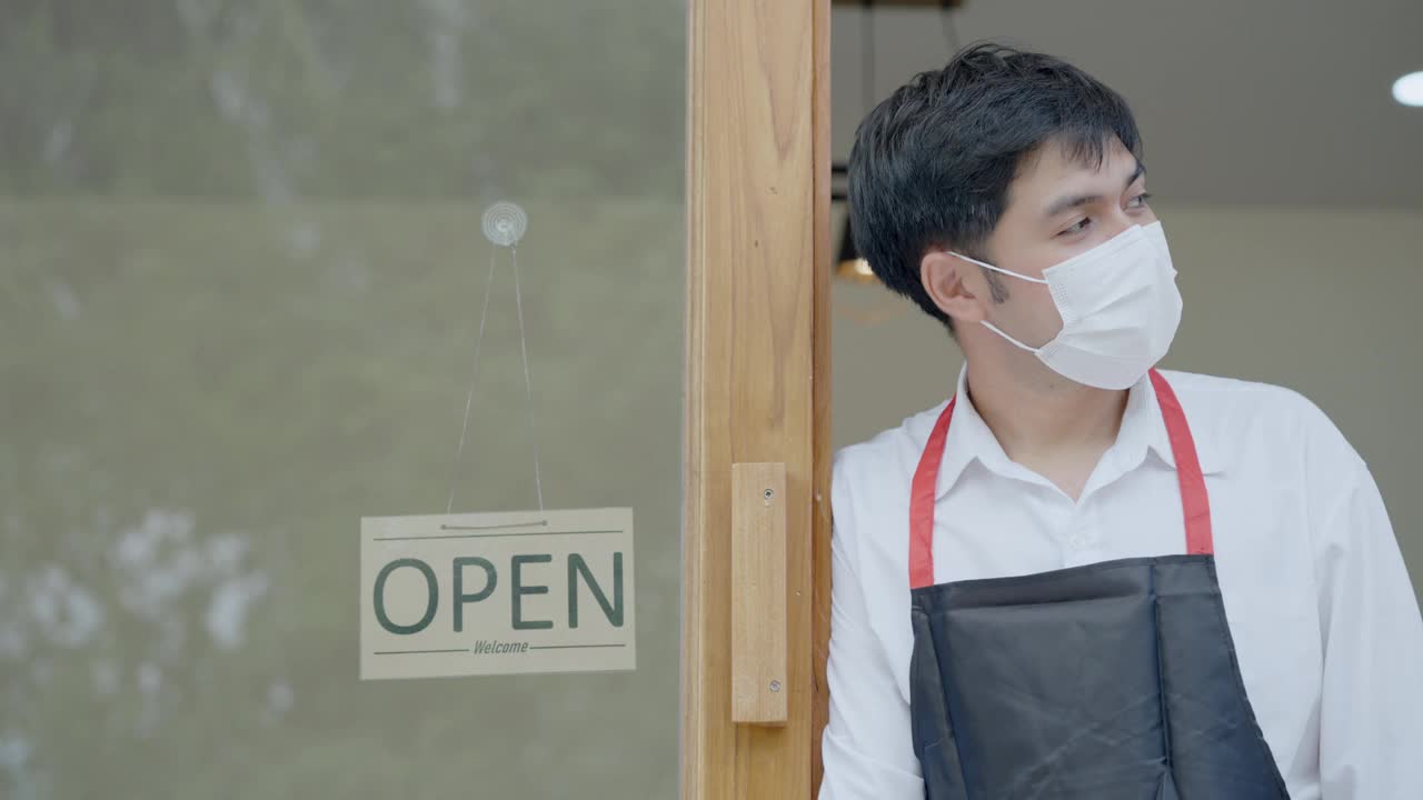 开业仪式的第一天，服务员戴着防护口罩，打开了咖啡店现代玻璃门上的招牌。新标准的概念。视频素材