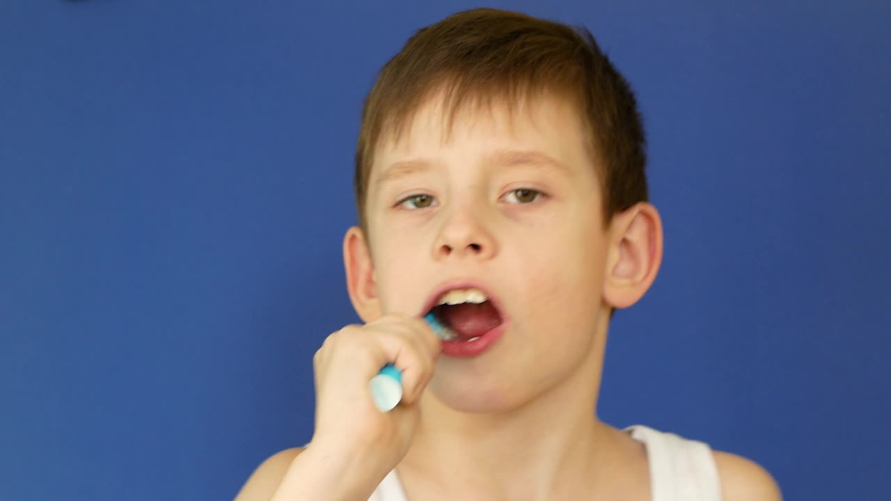 在摄影棚拍摄时，一个有魅力的8岁白人男孩一边刷牙一边唱歌，一边看着蓝色背景下的镜头。孩子早上在浴室里洗脸。牙科保健视频下载