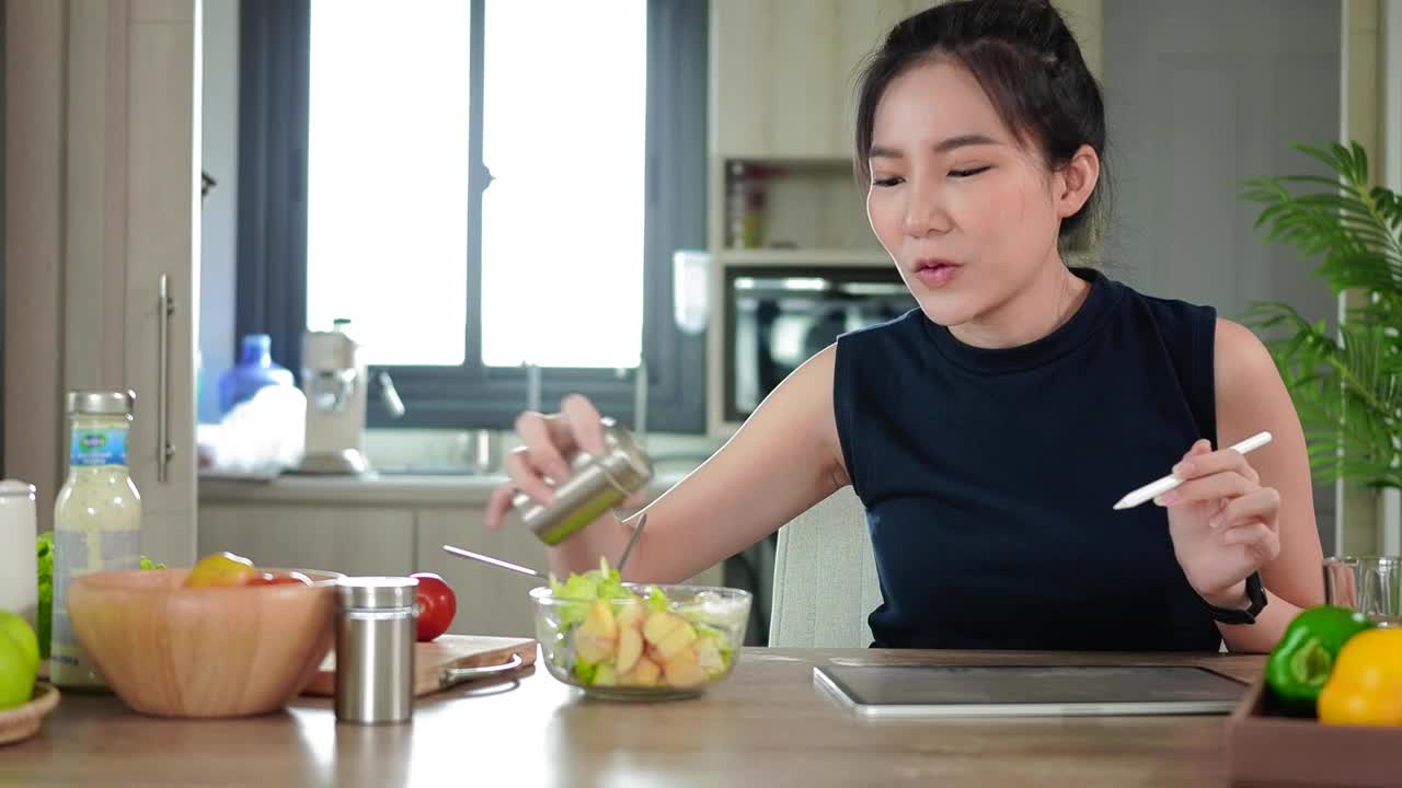快乐的亚洲女人在厨房用数码平板电脑吃新鲜的沙拉当早餐。视频素材