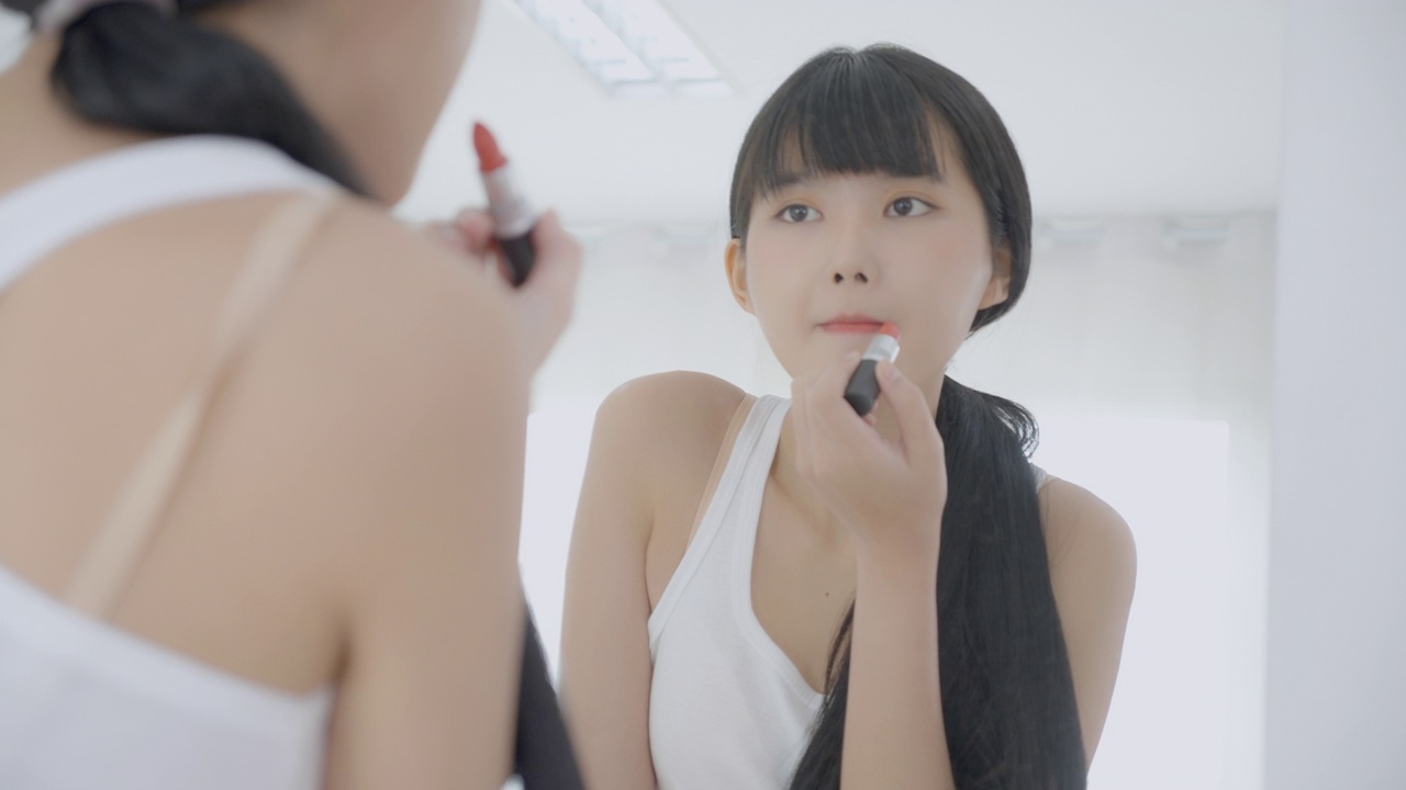 美丽年轻的亚洲女性涂口红在嘴上，美丽的女孩看着镜子的脸化妆化妆品与嘴唇的优雅，化妆与性感的女性，皮肤护理和健康的理念。视频下载