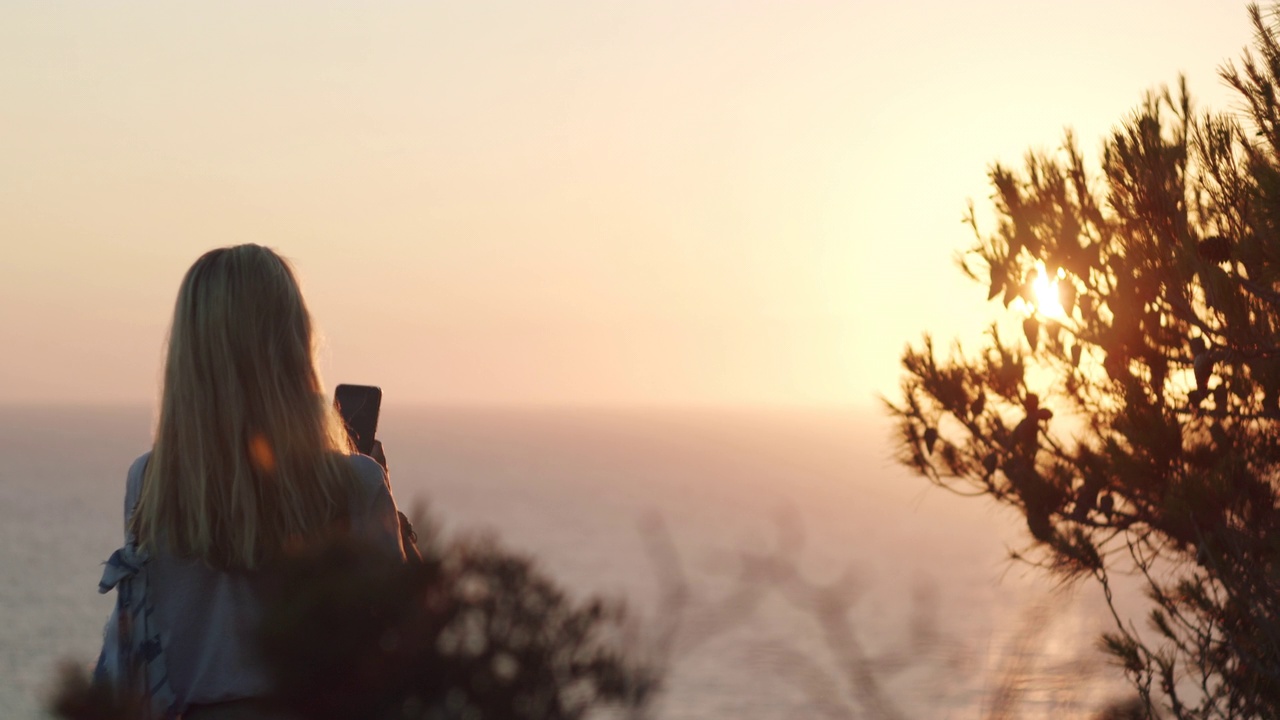 一个女人拿着手机，在海面上拍摄日落。暑假假期周末。视频素材