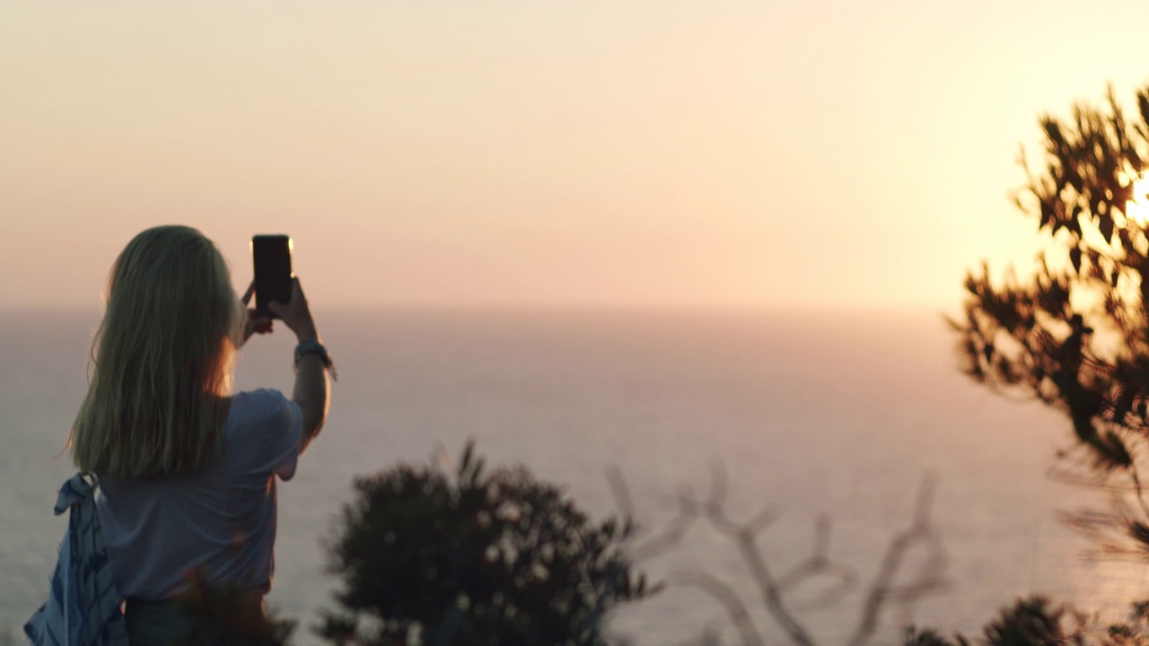一个女人拿着手机，在海面上拍摄日落。暑假假期周末。视频素材