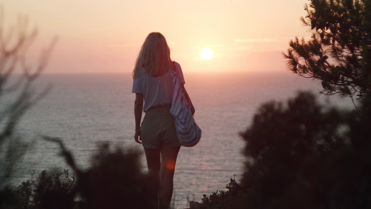 女人走向美丽的晚霞欣赏海景。暑假的假期概念。视频素材