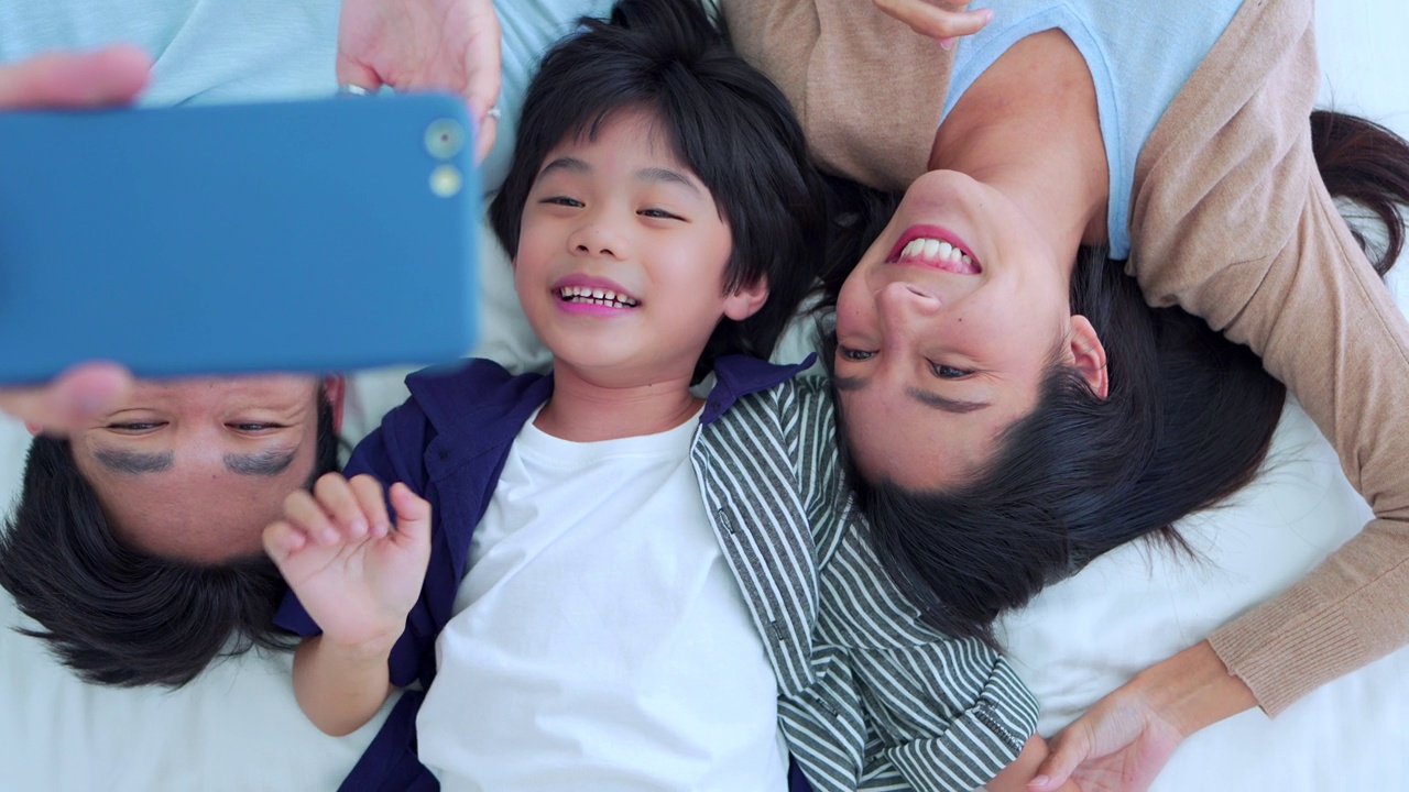 快乐的家庭时光，拿着自拍手机的亚洲父母爸爸，妈妈和儿子一起躺在床上的卧室里，开心的笑着。周末带孩子，享受在家放松的好时光。视频下载